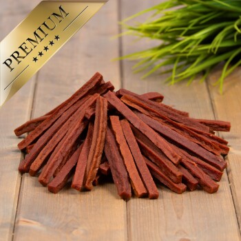 Ein Haufen hochwertiger Hundeleckerlis, die langen, dünnen Streifen ähneln, liegt auf einer Holzoberfläche mit grünem Gras im Hintergrund.