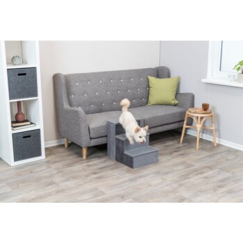 Ein kleiner weißer Hund klettert eine graue Hundetreppe neben einem grauen Sofa mit einem grünen Kissen in einem modernen Wohnzimmer.