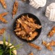 Getrocknete Fischleckerlis in einer schwarzen Schüssel, umgeben von verstreuten Stücken auf einer grauen Oberfläche, mit einer Pflanze und Seilspielzeugen in der Nähe.