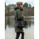 Eine Frau in einer grünen Parka und schwarzen Jeans steht an einem ruhigen See, trägt eine schwarze Tasche mit dem Etikett "WILD HAZEL."