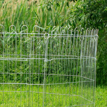 Drahtgehege für Haustiere im Freien auf grünem Gras mit hohen Pflanzen im Hintergrund.