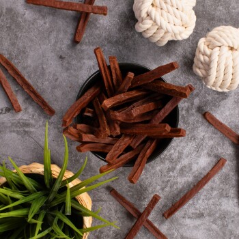Eine schwarze Schüssel gefüllt mit braunen Hundeleckerlis in Form von Stäbchen, umgeben von verstreuten Leckerlis, einer kleinen grünen Pflanze und zwei weißen Seilspielzeugen.