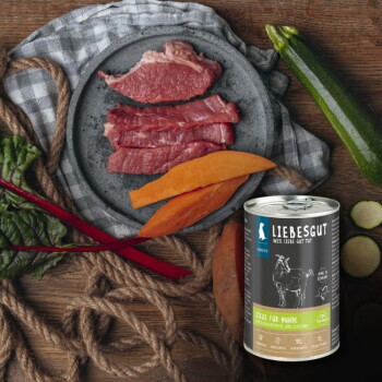 Dose Liebesgut-Hundefutter mit der Aufschrift 'Ziege für Hunde' neben rohem Fleisch, Süßkartoffelscheiben, Zucchini und Blattgemüse auf einem Holztisch.