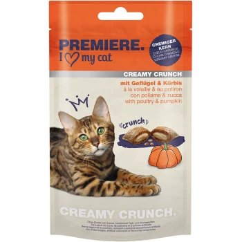 Eine Bengal-Katze mit grünen Augen ruht neben einer Packung "Premiere I Love My Cat Creamy Crunch" Leckerlis, die Geflügel und Kürbis enthält.