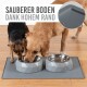 Zwei Hunde, ein brauner Golden Retriever und ein kleiner schwarz-brauner Hund, fressen aus Schalen auf einer grauen Matte mit dem Text: "SAUBERER BODEN DANK HOHEM RAND."