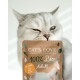 Eine Nahaufnahme einer grauen Katze, die verspielt ein Paket 'CAT'S LOVE 100% Bio Adult' Katzenfutter hält, mit Text auf Deutsch und Englisch.