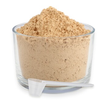 Eine Glas-Schüssel gefüllt mit hellbraunem Pulver, begleitet von einem kleinen weißen Messlöffel, der an der Schüssel lehnt.