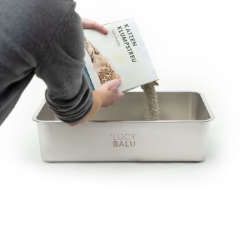 Eine Person in einem grauen Pullover schüttet Katzenstreu aus einer Box mit der Aufschrift "Katzen Klumpstreu" in eine Edelstahl-Katzenklo mit der Marke "LUCY BALU."