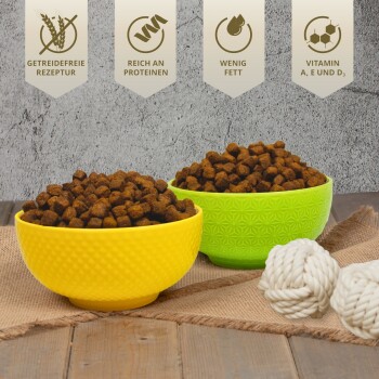 Zwei Schalen mit braunem Futter, eine gelbe und eine grüne, mit weißen Seilspielzeugen. Text: "Getreidefrei", "Hoher Proteingehalt", "Fettarm", "Vitamine A, E, D3."
