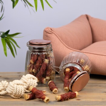 Ein gekipptes Glasgefäß mit Hundeleckerlis und gerollten Fleischstangen auf einem Holztisch, mit einem Seilspielzeug und einem rosa Haustierbett im Hintergrund.