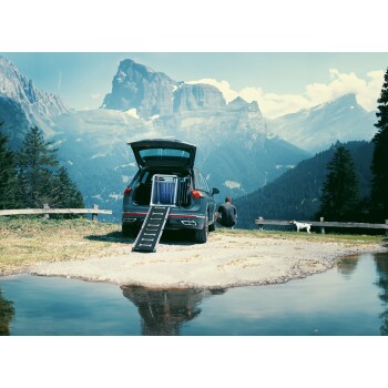 Ein Mann sitzt neben einem Auto mit offenem Kofferraum, in dem eine Haustierrampe zu sehen ist, in einer malerischen Berglandschaft mit einem kleinen Hund in der Nähe.