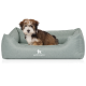 Ein kleiner, flauschiger Welpe mit braunem und weißem Fell sitzt bequem in einem hellgrünen Hundebett, das das Logo "KNUFFELWUFF" trägt.