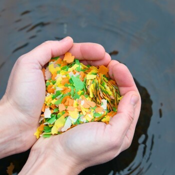 Die Hände einer Person, die bunte Fischfutterflocken in den Farben Grün, Orange und Weiß halten, mit einer verschwommenen Wasseroberfläche im Hintergrund.