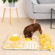 Ein kleiner brauner Hund interagiert mit einer bunten, interaktiven Haustierpuzzle-Matte, die verschiedene Texturen und Formen in Gelb und Beige aufweist.