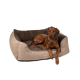 Ein brauner Hund liegt auf einem plüschigen, beigen Hundebett mit einem dunkelbraunen Innenraum, auf dessen Seite der Text 'TrendPet' gestickt ist.