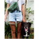 Eine Frau in einem grauen T-Shirt und Jeansshorts hält eine gemusterte Hundeleine, steht neben einem lächelnden schwarzen Hund mit braunen Augen.