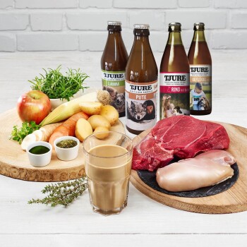 Frische Zutaten für Haustierfutter: rohes Rind- und Hühnerfleisch, Gemüse, ein Apfel, Kräuter und vier Tjure-Getränke mit den Bezeichnungen "Lamm," "Pute," "Rind," "Geflügel."