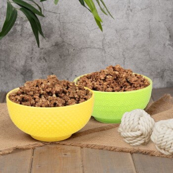 Zwei bunte Schalen, eine gelbe und eine grüne, gefüllt mit Haustierfutter, stehen auf einer Juteschicht neben zwei weißen Seilspielzeugen.