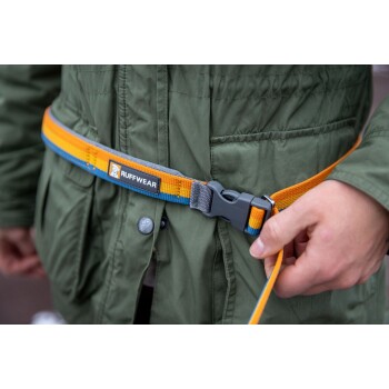 Eine Person passt einen bunten Ruffwear-Hundeleinen-Gürtel um ihre Taille an, der orangefarbene, blaue und graue Riemen hat, während sie eine grüne Jacke trägt.