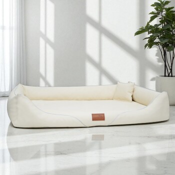 Cremefarbenes rechteckiges Haustierbett mit erhöhten Seiten und kleinem Kissen, auf Marmorboden neben einer Pflanze platziert.