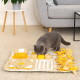 Graue Katze untersucht ein gelb-beiges Schnüffelmatten-Spielzeug auf cremefarbenem Teppich neben einem grauen Sofa und einem Tisch mit Pflanze.
