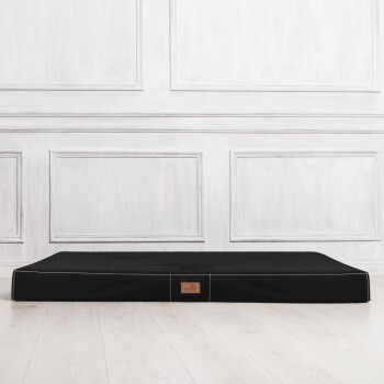 Schwarzes Haustierbett mit einem Lederetikett, vor einer weißen Paneelwand und einem hellen Holzboden.