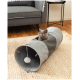 Eine graue Katze mit gelben Augen sitzt in einem grauen und schwarzen Stofftunnelspielzeug, das einen baumelnden Pom-Pom hat, auf einem gewebten Teppich.