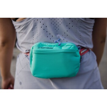 Eine Nahaufnahme einer Person in einem hellgrauen Sportoutfit, mit einer leuchtend türkisfarbenen Ruffwear-Tasche, die an ihrer Taille befestigt ist.