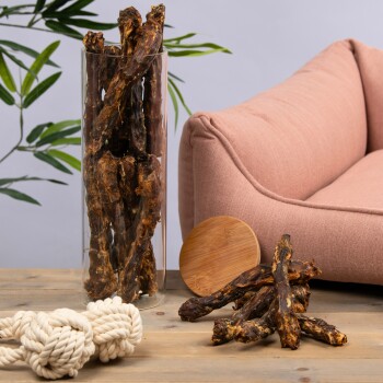 Eine Glasvase, gefüllt mit Hundekauartikeln, steht neben einem rosa Haustiersofa, mit einem Holzbrett und einem weißen Seilspielzeug auf einem Holztisch.