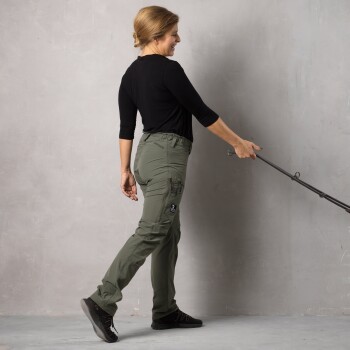 Eine Frau in einem schwarzen Langarmshirt und olivgrünen Cargo-Hosen geht mit einer Hundeleine in der rechten Hand, vor einem grauen Hintergrund.