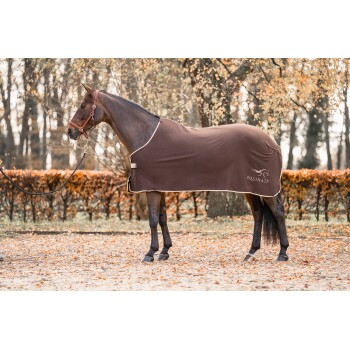 Ein braunes Pferd, das eine braune Decke mit dem Logo "EQUINATE" trägt, steht in einer mit Blättern bedeckten Umgebung, umgeben von Bäumen.