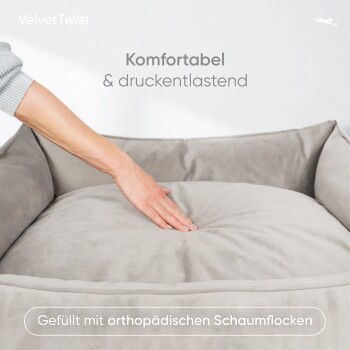 Eine Person drückt ein plüschiges graues Haustierbett mit Textüberlagerung: "Komfortabel & druckentlastend" und "Gefüllt mit orthopädischen Schaumflocken."