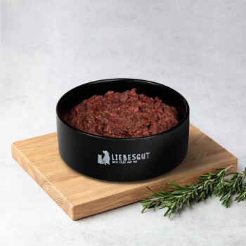 Schwarzer Napf mit Aufschrift 'LIEBESGUT WEIL LIEBE GUT TUT', gefüllt mit rohem Hackfleisch, auf einem Holzbrett mit Rosmarin