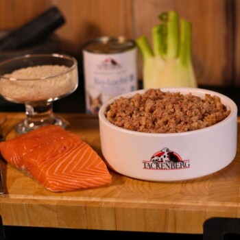 Biała miska z mieloną karmą dla zwierząt oznaczona "Tackenberg" z surowym łososiem, ryżem, koprem włoskim oraz puszka karmy dla zwierząt w tle.