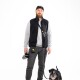 Eine Person in einer schwarzen Weste und einem grauen Pullover steht mit einem kleinen schwarzen Hund an der Leine und hält eine graue Wasserflasche.