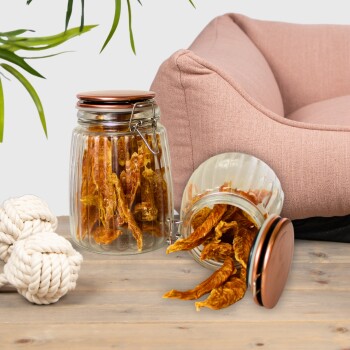 Ein Glasgefäß gefüllt mit getrockneten Haustierleckereien, das auf eine Holzoberfläche neben einem plüschigen rosa Haustierbett und einem Seilspielzeug kippt.