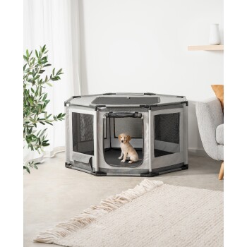 Ein kleiner Welpe sitzt in einem grauen Haustierspielzaun mit Netzseiten, der auf einem hellen Boden neben einem gemütlichen Sofa und einer Pflanze platziert ist.