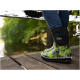 Grün-schwarz gemusterte Gummistiefel, die von einer Person auf einem Holzsteg an einem ruhigen, bewaldeten Wasserlauf getragen werden.