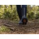 Eine Person in schwarzen Gummistiefeln geht auf einem Waldweg, der mit Kiefernnadeln und Moos bedeckt ist, mit einem sichtbaren Tannenzapfen auf dem Boden.