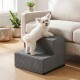 Kleiner weißer, flauschiger Hund auf grauer Haustiertreppe mit zwei Stufen, lehnt an einem beigen Sofa in einem sonnendurchfluteten Wohnzimmer mit Pflanzen.