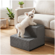 Kleiner weißer, flauschiger Hund auf grauer Haustiertreppe mit zwei Stufen, lehnt an einem beigen Sofa in einem sonnendurchfluteten Wohnzimmer mit Pflanzen.
