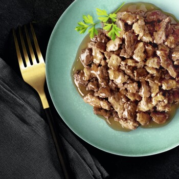 Close-up van natte huisdiervoeding met vleesstukjes in jus, gegarneerd met peterselie, naast een gouden vork op een donkere achtergrond.