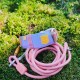 Ein farbenfrohes Hundegeschirr in Lila und Braun, kombiniert mit einer rosa Leine, die auf grünem Moos ruht. Das Geschirr hat ein gelbes Logo-Patch.