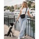 Lächelnde Frau in weißem Crop-Top und Jeans, die eine Leine und eine schwarze Umhängetasche hält, mit einem schwarz-weißen Border Collie an einer Marina; kleiner weißer vertikaler Text rechts.