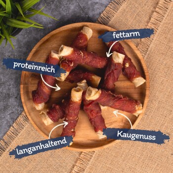 Ein Holztablett zeigt gerollte Hundeleckerlis mit den Bezeichnungen "hoher Proteinanteil", "fettarm", "langanhaltend" und "Kaugenuss."