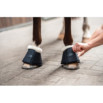 Eine Person befestigt einen schwarzen Pferdestiefel mit einem flauschigen weißen Innenfutter um das Vorderbein eines Pferdes und zeigt das Logo des Stiefels.