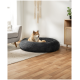 Ein runder grauer Hundebett auf einem Holzboden, in dem ein ruhender Hund liegt, mit einem hellen Teppich und einem modernen Sofa im Hintergrund.