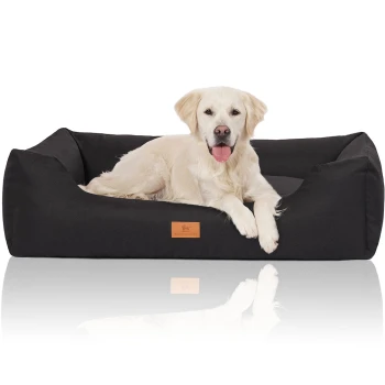 Hundebett Lotte aus Velours mit feinem Handwebcharakter schwarz XL Ein Golden Retriever liegt bequem auf einem schwarzen Hundebett mit einem Holzschild mit der Aufschrift "HUTTEWOR." Das Bett hat ein modernes Design.