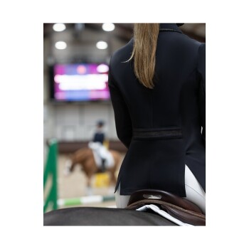 Ein Reiter in einer schwarzen Reitjacke sitzt auf einem Pferd, mit einem verschwommenen Hintergrund einer Springarena und einem anderen Teilnehmer.