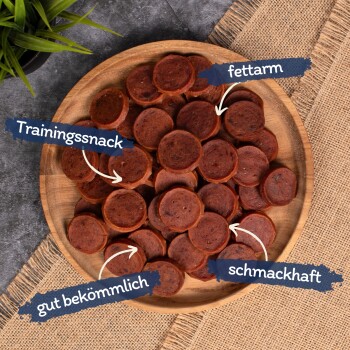 Ein Holzbrett mit runden, geschnittenen Hundetraining-Leckerlis, beschriftet auf Deutsch: "Trainingssnack", "fettarm", "gut bekömmlich" und "schmackhaft."
